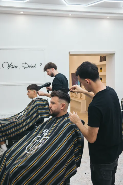 Barber upravuje strih klientovi s bradou, v pozadí ďalší barber pri práci